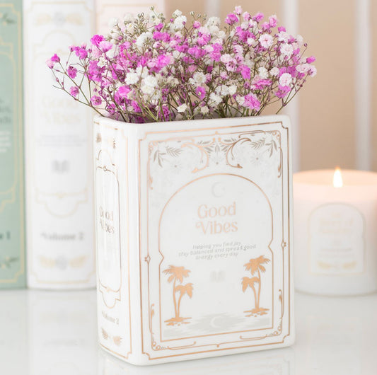 Book shaped vase. White with gold delicate details including the words ‘good vibes’. Book is standing upright with flowers inserted inside where the pages would be. 
